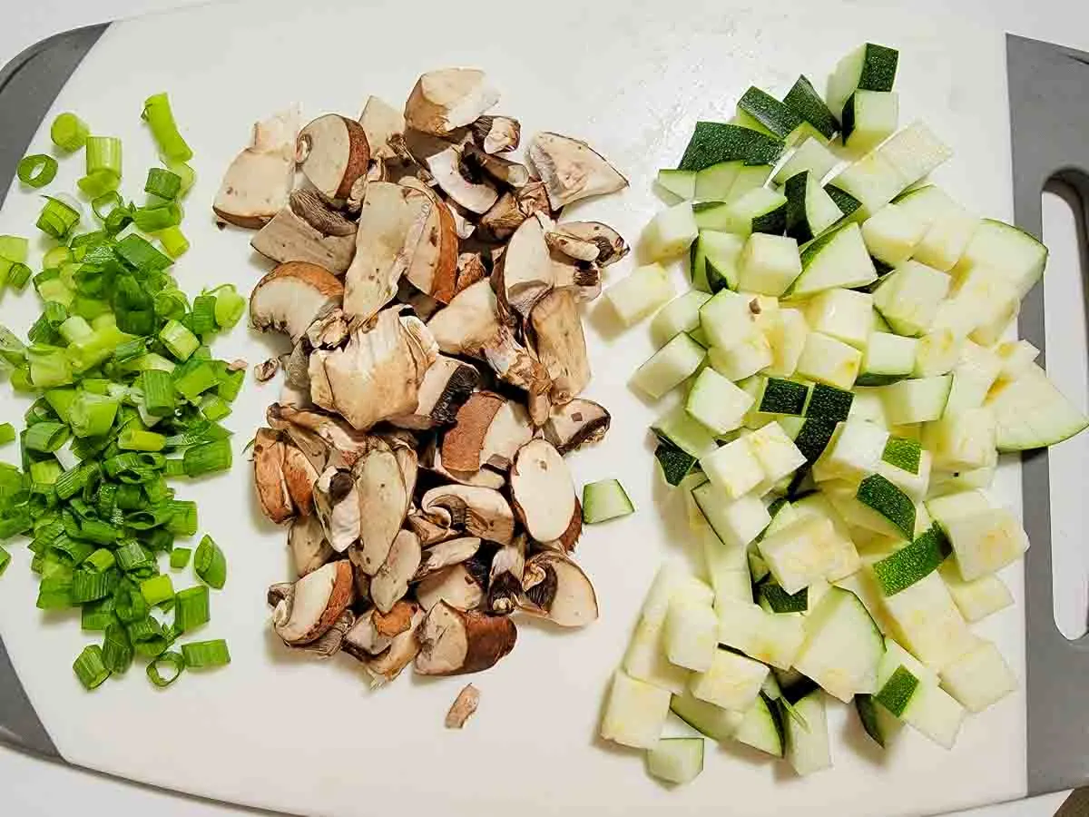 green onions, mushrooms, and zucchini diced on a cutting board.
