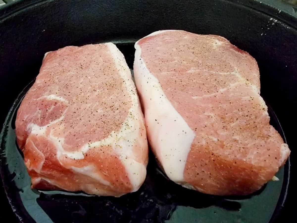 two pork chops cooking in a cast iron skillet.