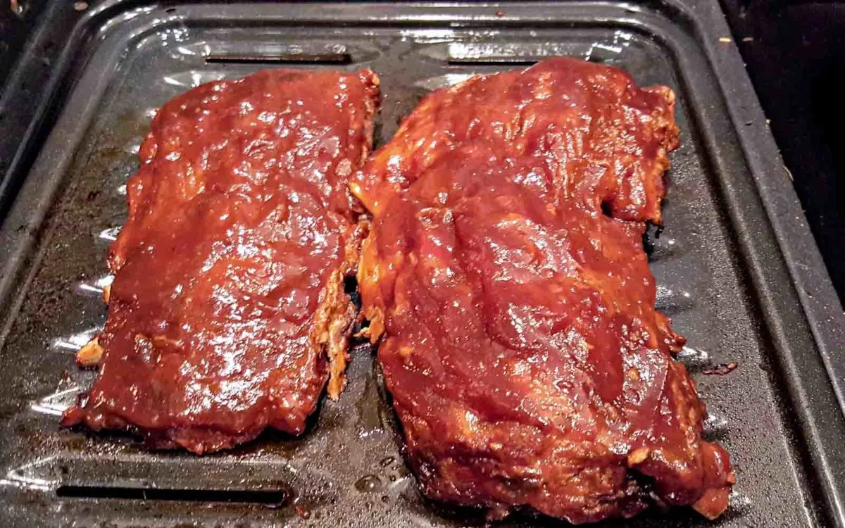 two slabs of baby back ribs covered in barbecue sauce on a broiler pan.