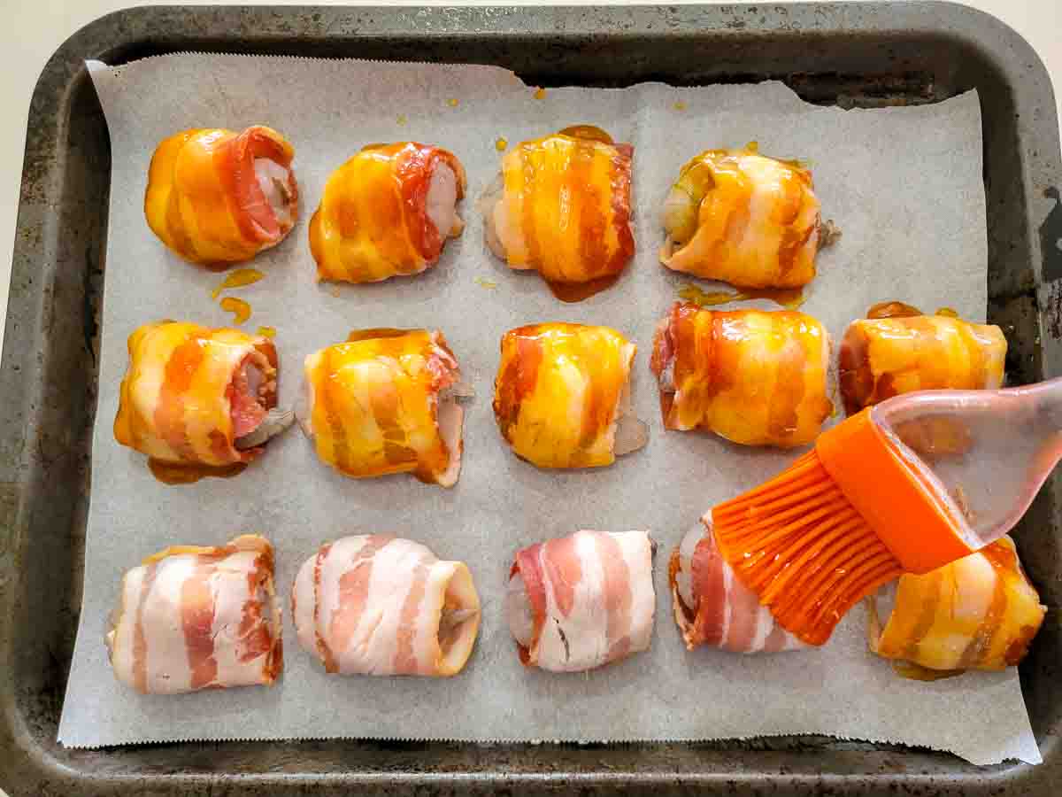 brushing apricot jam on raw shrimp topped with a mozzarella ball and wrapped in a slice of bacon on a cutting board.