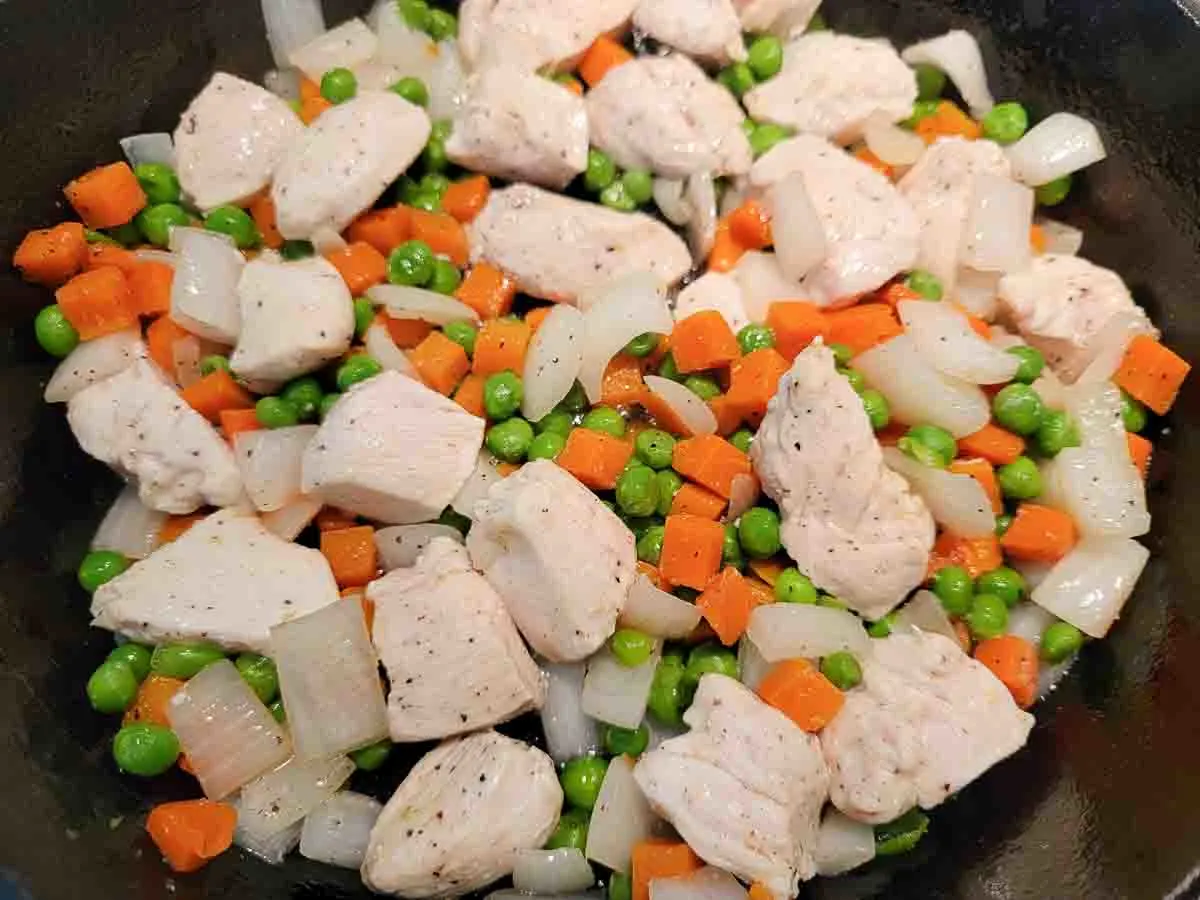 chicken, peas, carrots, and onions cooking in a cast iron skillet.