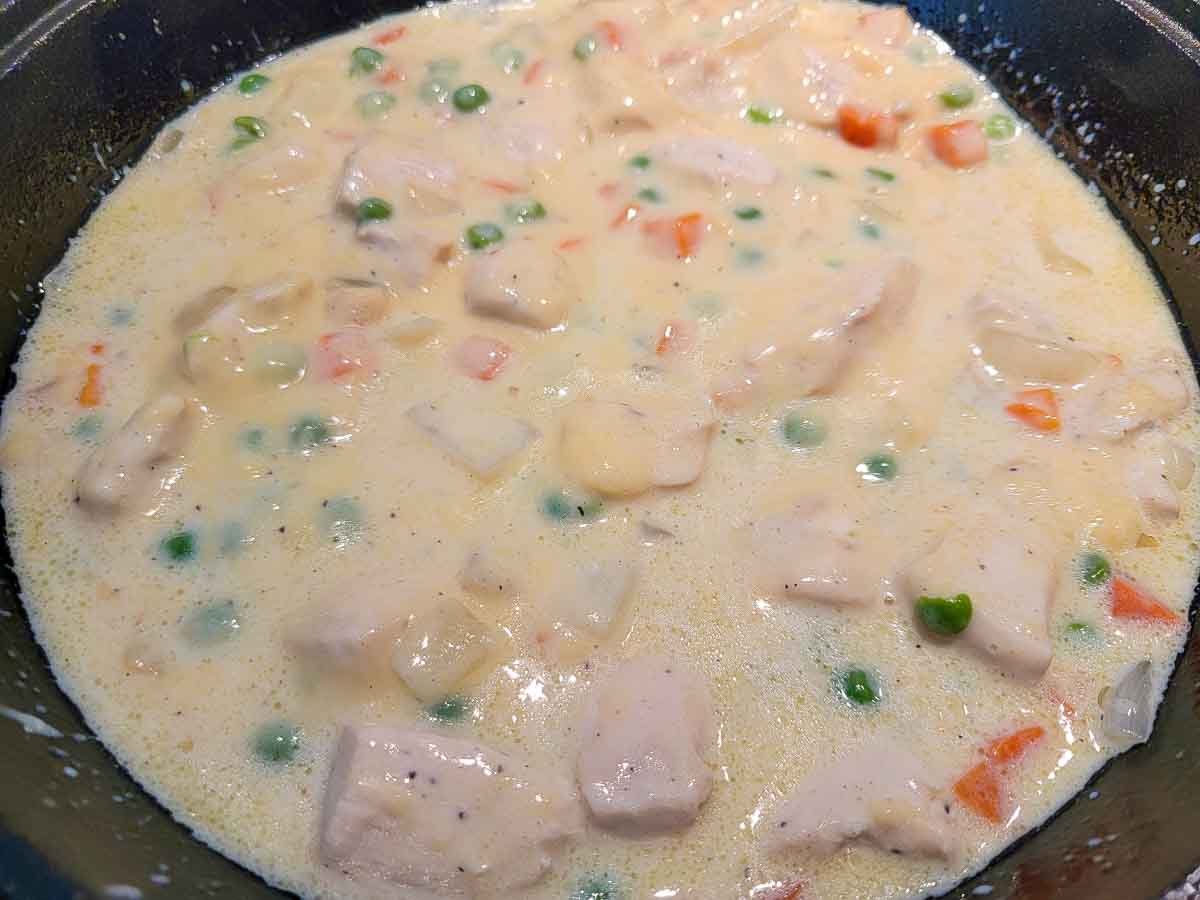 cream of chicken soup, milk, salt, pepper, chicken, peas, carrots, and onions cooking in a cast iron skillet.