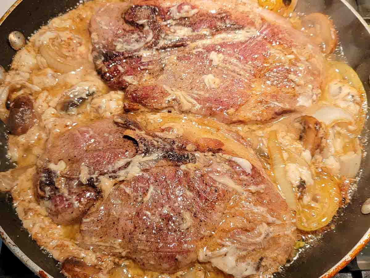 pork steaks, garlic, cream of mushroom soup, onion slices and mushroom slices cooking in a pan.