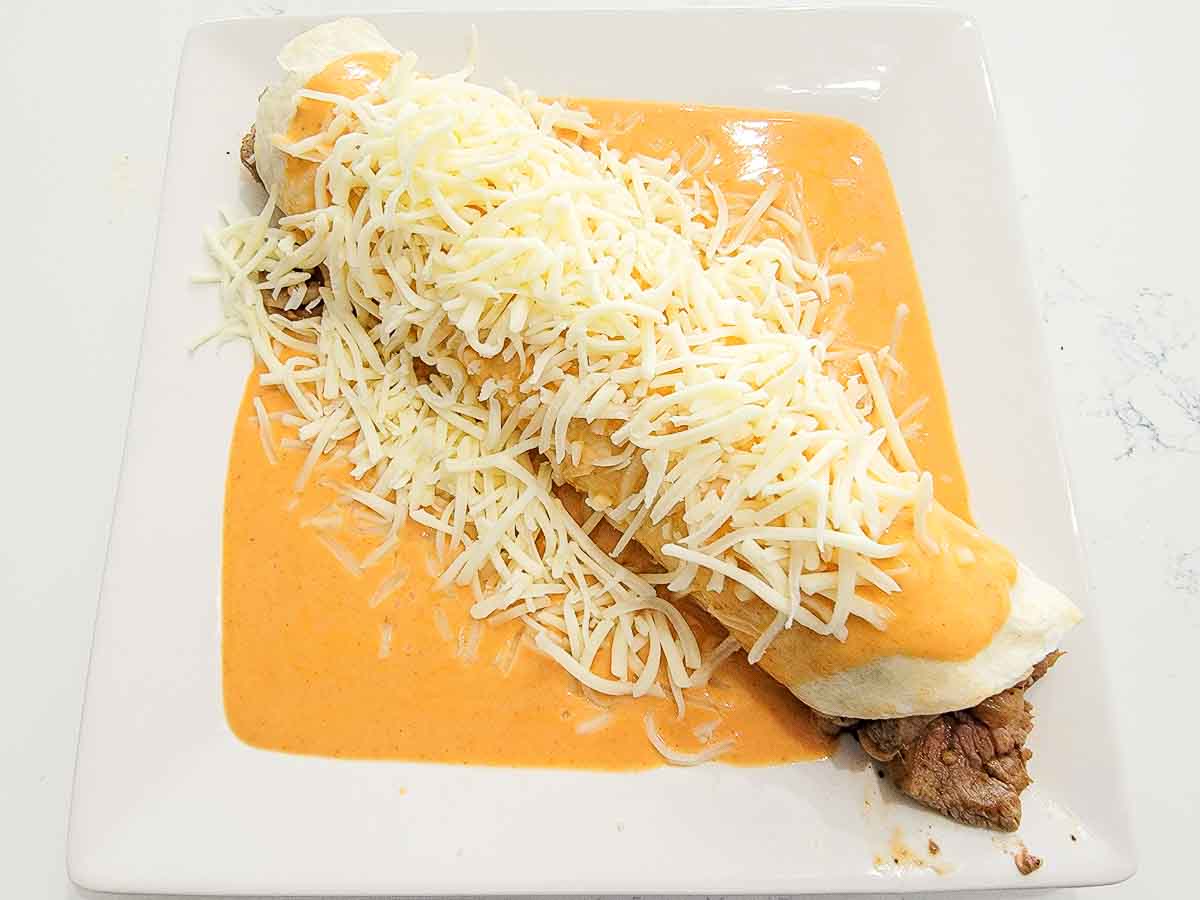 a steak burrito on a plate covered in orange sauce and shredded cheese.