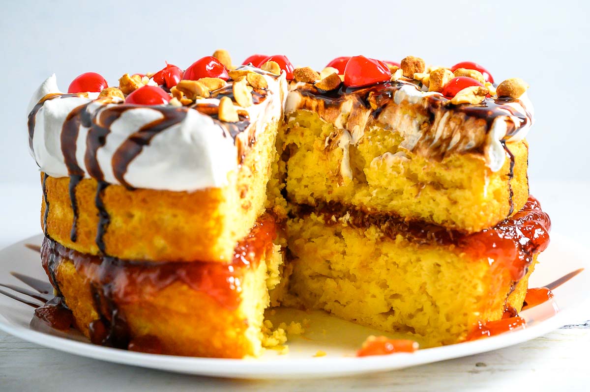 a cake with layers of yellow cake mix, banana pudding, crushed pineapple, strawberry jam, cool whip, chocolate syrup, and maraschino cherries.