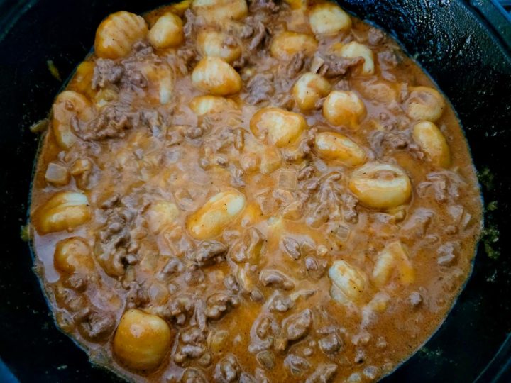 cheddar cheese, ground beef, onions, salt, garlic powder, paprika, chili powder, tomato paste, Worcestershire sauce, heavy cream, beef broth, gnocchi, and pepper cooking in a cast iron skillet.