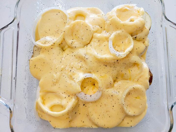 potato slices and onion slices topped with black pepper and cheese sauce in a baking dish.